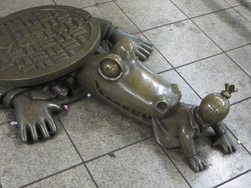 Alligator Eats Moneybags by edenpictures portrays a NYC subway alligator sculpture popping out of a manhole in order to eat a sculpture of a person with a money bag as a head